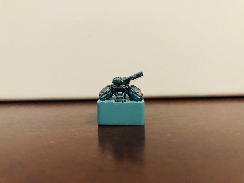May include: A small, gray, and black miniature tank with a red stripe on the side, sitting on a gray base.