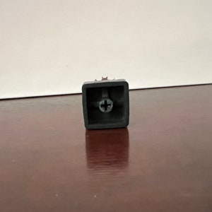 Puede incluir: Un tap&oacute;n de tecla de pl&aacute;stico negro con un tornillo plateado en el centro.