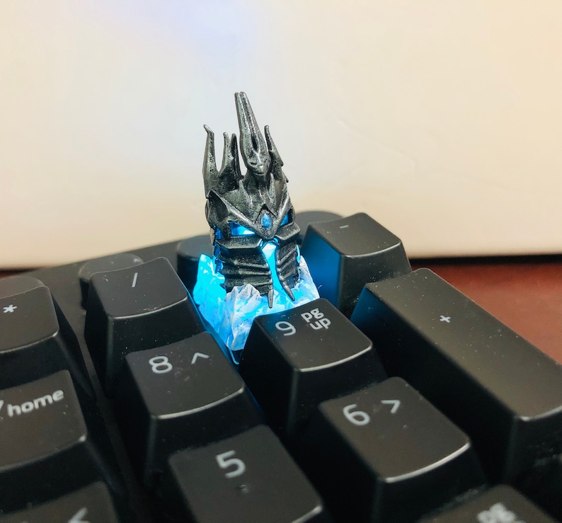 May include: A black and silver 3D printed keycap featuring a crown with blue glowing lights. The keycap is placed on a black keyboard with the keys labeled "8", "9", "Pg Up", "6", and "5".
