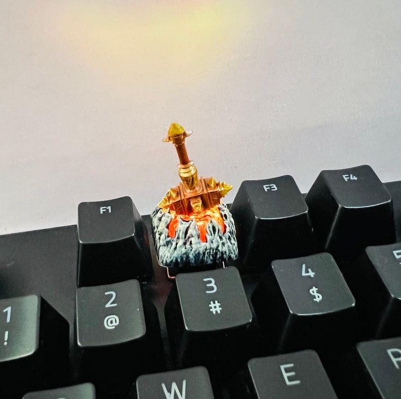 Op de afbeelding: Een gouden miniatuurzwaard met een zwart-witte basis, dat op een zwart toetsenbord ligt met toetsen met de labels 'F1', 'F3', 'F4', '2', '3', '4', 'E', 'W', 'P' en 'F'.