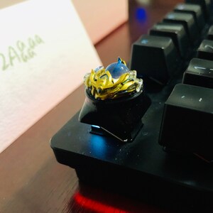 Puede incluir: Un tap&oacute;n de tecla dorado y azul con un dise&ntilde;o de corona y una gema azul en el centro. El tap&oacute;n de tecla est&aacute; en un teclado negro.