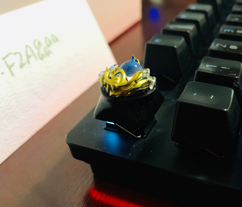 Puede incluir: Un tap&oacute;n de tecla dorado y azul con un dise&ntilde;o de corona y una gema azul en el centro. El tap&oacute;n de tecla est&aacute; en un teclado negro.