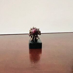 Puede incluir: Una peque&ntilde;a escultura detallada de bronce de una criatura con un gran ojo morado oscuro. La escultura est&aacute; sobre una base negra.