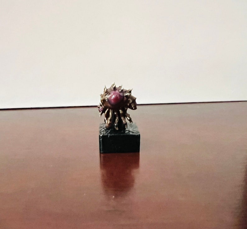 Puede incluir: Una peque&ntilde;a escultura detallada de bronce de una criatura con un gran ojo morado oscuro. La escultura est&aacute; sobre una base negra.