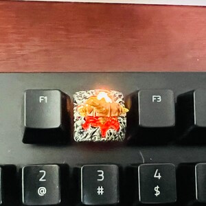 Op de afbeelding: Een close-up van een aangepaste toetsenbord-keycap met een gedetailleerd ontwerp in rood, wit en goud. De keycap wordt van onderen verlicht, waardoor een warme gloed ontstaat.