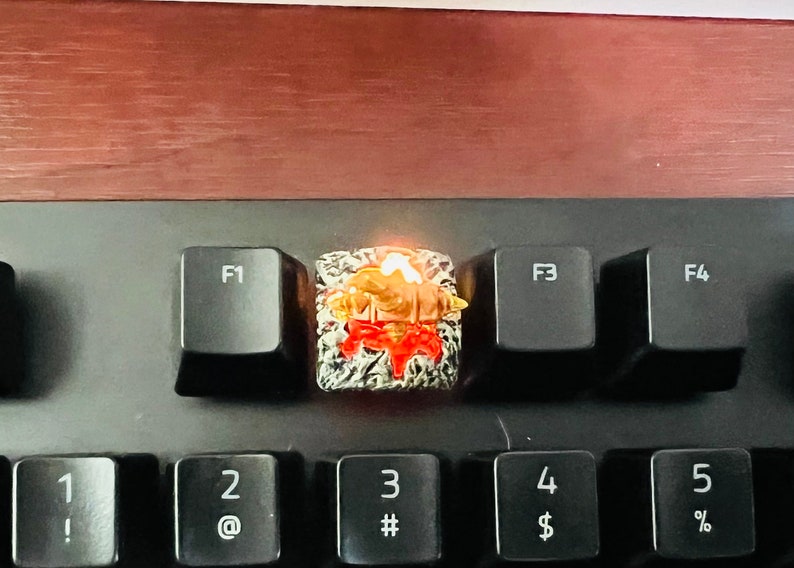 Op de afbeelding: Een close-up van een aangepaste toetsenbord-keycap met een gedetailleerd ontwerp in rood, wit en goud. De keycap wordt van onderen verlicht, waardoor een warme gloed ontstaat.