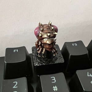 Puede incluir: Un tap&oacute;n de tecla impreso en 3D que presenta una criatura detallada de color bronce con armadura puntiaguda y grandes ojos rosados. La criatura est&aacute; posada sobre una base negra y est&aacute; dise&ntilde;ada para adaptarse a un teclado est&aacute;ndar.