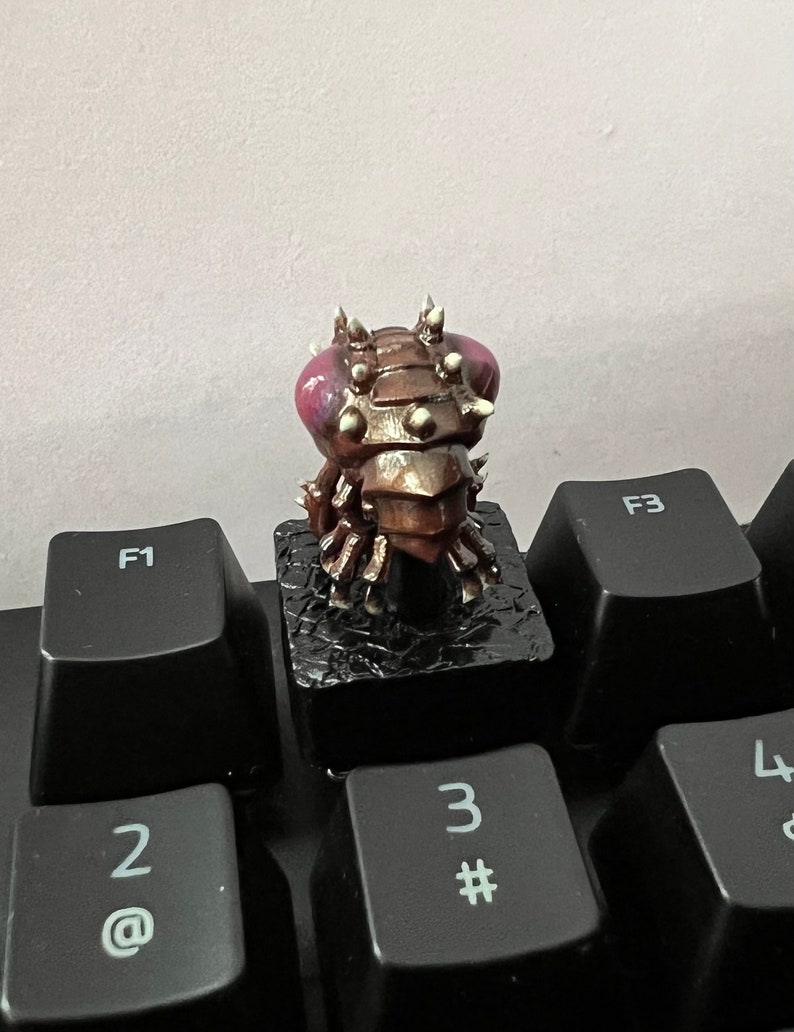 Puede incluir: Un tap&oacute;n de tecla impreso en 3D que presenta una criatura detallada de color bronce con armadura puntiaguda y grandes ojos rosados. La criatura est&aacute; posada sobre una base negra y est&aacute; dise&ntilde;ada para adaptarse a un teclado est&aacute;ndar.