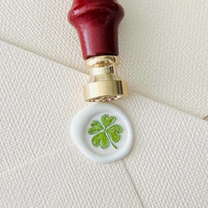 Op de afbeelding: Een witte waszegel met een groen vierbladig klavertjevier. De zegel is bevestigd aan een witte envelop. Een gouden en rode waszegel stempel bevindt zich boven de zegel.
