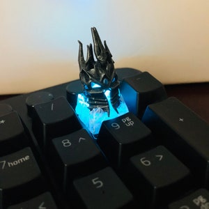 May include: A black and silver 3D printed keycap featuring a detailed crown design. The keycap is lit from below with a blue light, illuminating the intricate details of the crown.