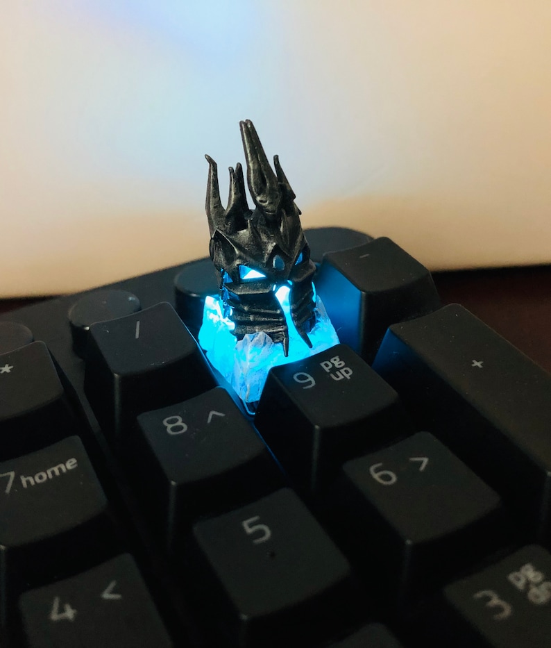 May include: A black and silver 3D printed keycap featuring a detailed crown design. The keycap is lit from below with a blue light, illuminating the intricate details of the crown.