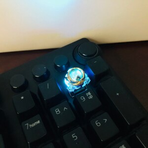 Może przedstawiać: Czarna klawiatura z podświetlanym niebieskim klawiszem. Klawisz ma złoty pierścień i jest oznaczony cyfrą "9" i tekstem "pg up".