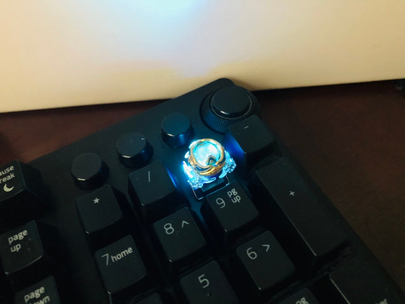 Może przedstawiać: Czarna klawiatura z podświetlanym niebieskim klawiszem. Klawisz ma złoty pierścień i jest oznaczony cyfrą "9" i tekstem "pg up".