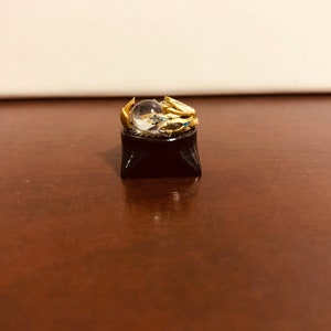 Puede incluir: Un tap&oacute;n de tecla dorado con una esfera de cristal transparente en la parte superior, sentado sobre una base de tecla negra.