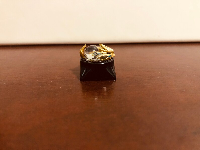 Puede incluir: Un tap&oacute;n de tecla dorado con una esfera de cristal transparente en la parte superior, sentado sobre una base de tecla negra.