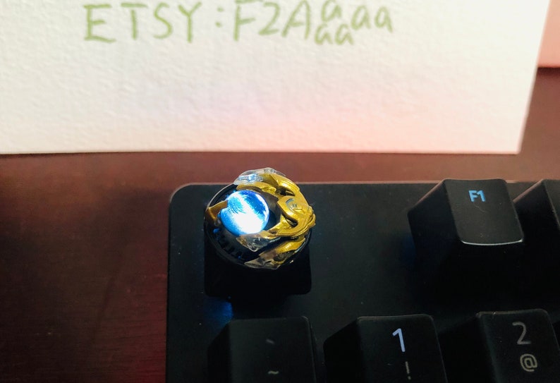 Puede incluir: Un tap&oacute;n de tecla dorado y azul con una luz azul brillante en su interior. El tap&oacute;n de tecla est&aacute; en un teclado negro.