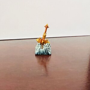Op de afbeelding: Een miniatuur gouden hamer met spikes en een bruine steel, gemonteerd op een grijze en witte rotsbasis.
