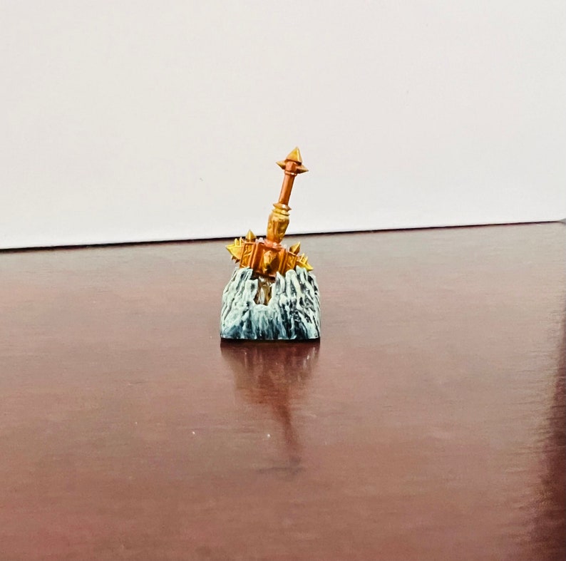 Op de afbeelding: Een miniatuur gouden hamer met spikes en een bruine steel, gemonteerd op een grijze en witte rotsbasis.