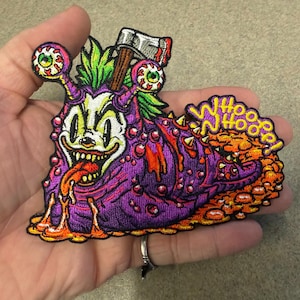 May include: An embroidered patch featuring a purple slug-like creature with a clown face, green hair, and an axe embedded in its head. The creature has two eyes on stalks, a long tongue, and the yellow text WHOOOO NHOOOO! on an orange trail.