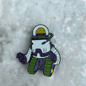 May include: A green, purple, and white enamel pin of a robot with a yellow brain inside a clear dome. The robot has a purple body, green legs, and a white head.