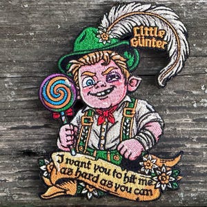 May include: An embroidered patch featuring a character wearing a green hat with a white feather and the text Little Günter. The character holds a colorful spiral lollipop and wears suspenders. A banner below reads I want you to hit me as hard as you can.
