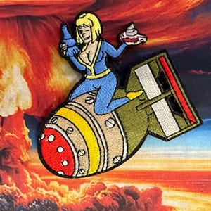 May include: Embroidered patch featuring a cartoon woman in a blue jumpsuit, riding a bomb. She holds a bottle and a cupcake. The bomb is olive green with red and yellow accents. The background shows a nuclear explosion.