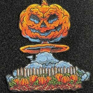May include: Embroidered patch featuring a grinning jack-o'-lantern with an orange and blue color scheme. Below the pumpkin is a mushroom cloud, a white picket fence, and pumpkins. The patch is on a black background.