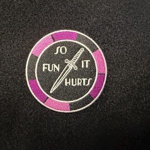 May include: A circular embroidered patch featuring a black center with a white dagger graphic and the text SO FUN IT HURTS. The border consists of alternating black and magenta segments, finished with a white stitched edge on a black background.