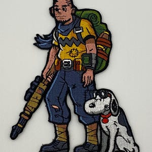 May include: Embroidered patch featuring a cartoon character with a backpack, rifle, and a dog wearing goggles. The character wears a yellow shirt, blue pants, and boots. The dog is white with black spots and a red collar. The patch has a white background.
