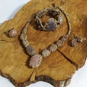 Op de afbeelding: Een set sieraden met een ketting, armband en oorbellen. De sieraden zijn gemaakt met natuurlijke stenen en macramé. De stenen zijn lichtroze en paars van kleur. Het macramé is van natuurlijke bruine kleur.