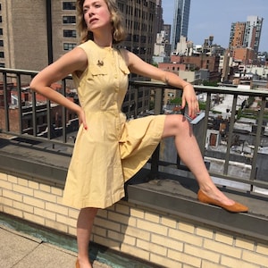 May include: A woman wearing a yellow sleeveless dress with a button-up front and a small gold pin on the chest. She is standing on a rooftop with a city skyline in the background.