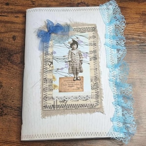May include: A white journal with a blue lace trim and a decorative cover featuring a vintage photo of a young girl in a plaid dress, a blue bow, and a floral background. The cover also includes a piece of burlap fabric and a small paper label with the text "PRIMAVERA - TRULY NATURAL COLORS".