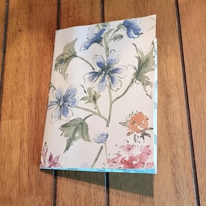 May include: A folded notebook with a floral design featuring blue, pink, and orange flowers on a cream background. The notebook is resting on a wooden surface.