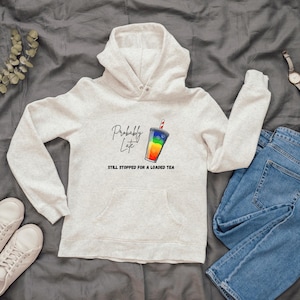 May include: A light gray hoodie with the text "Probably Late" and "Still Stopped for a Loaded Tea" printed on the front. The graphic is a rainbow-colored drink. The hoodie is paired with blue jeans and white sneakers.