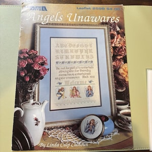 Op de afbeelding: Een ingelijst kruissteekpatroon getiteld "Angels Unawares" met een engelenmotief. Het patroon bevat het alfabet, een bijbelvers en engelenillustraties. Een theepot, bloemen en een kleine ronde engelenafbeelding zijn ook in de afbeelding te zien.