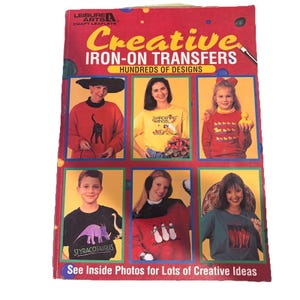 Könnte beinhalten: Ein Vintage-Buch mit dem Titel "Creative Iron-On Transfers" mit rotem Einband. Das Cover zeigt Bilder von Personen, die Hemden mit verschiedenen Designs tragen. Der Text "Hundreds of Designs" ist ebenfalls sichtbar. Das Buch ist ein Bastelheft.
