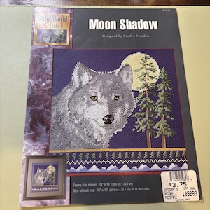 May include: A cross-stitch kit titled "Moon Shadow" featuring a detailed image of a wolf under a full moon and pine trees. The kit includes a framed image size of 14" x 14" (35.6 cm x 35.6 cm). Designed by Sandra Paradise.