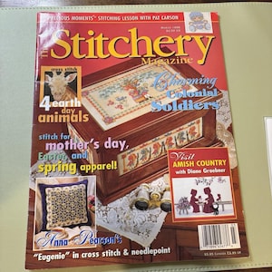 May include: The Stitchery Magazine cover features a wooden box with cross-stitch designs, a giraffe, and the text "Charming Colonial Soldiers." Additional text includes "4 earth day animals" and "stitch for mother's day, Easter, and spring apparel!"