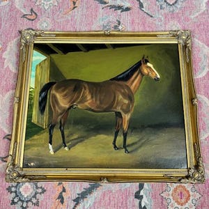 May include: An oil painting of a brown horse in a stable, framed in a gold ornate frame. The horse has a black mane and tail, with white markings on its face and legs. The background is a muted green and blue.