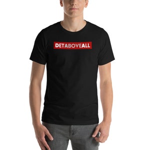 May include: Black t-shirt with the text "DETABOVEALL" in red letters inside a white rectangle. The shirt is short-sleeved and appears to be made of a soft material. The model is wearing grey trousers.