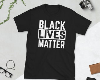 Może przedstawiać: Czarna koszulka z białym napisem "BLACK LIVES MATTER" w postarzanym kroju pisma, ułożonym w trzech liniach w białym prostokącie. Koszulka jest prezentowana na neutralnym tle.
