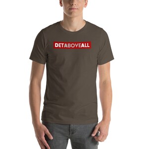 May include: A brown t-shirt with short sleeves, featuring the text "DETABOVEALL" in white within a red rectangle. The t-shirt is made of a soft material.