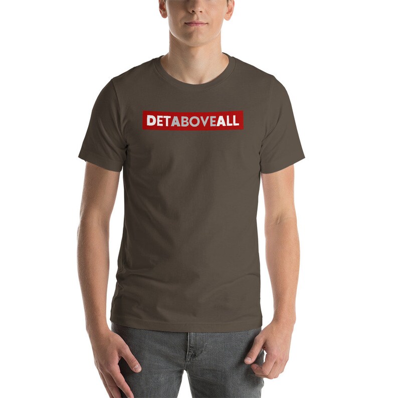 May include: A brown t-shirt with short sleeves, featuring the text "DETABOVEALL" in white within a red rectangle. The t-shirt is made of a soft material.