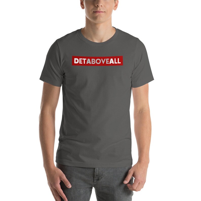 May include: A dark grey t-shirt featuring the text "DETABOVEALL" in white within a red rectangular box. The shirt has short sleeves and is likely made from a soft fabric.
