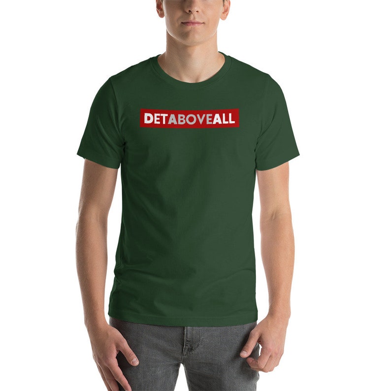 May include: A dark green t-shirt featuring the text "DETABOVEALL" in white letters within a red rectangle. The short-sleeved shirt is made of a soft material. The model is wearing grey trousers.