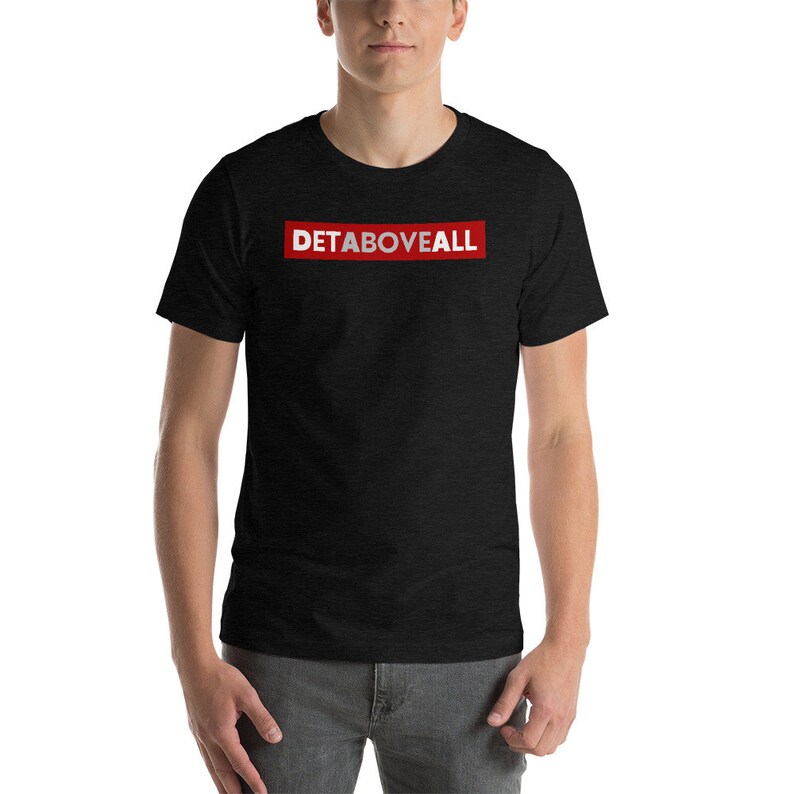 May include: Black t-shirt with the text "DETABOVEALL" in white letters inside a red rectangle. The shirt is short-sleeved and made of a soft material. The person is wearing grey trousers.