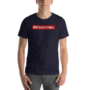 May include: A navy blue t-shirt featuring the text "DETABOVEALL" in white within a red rectangular box. The shirt has a crew neck and short sleeves.