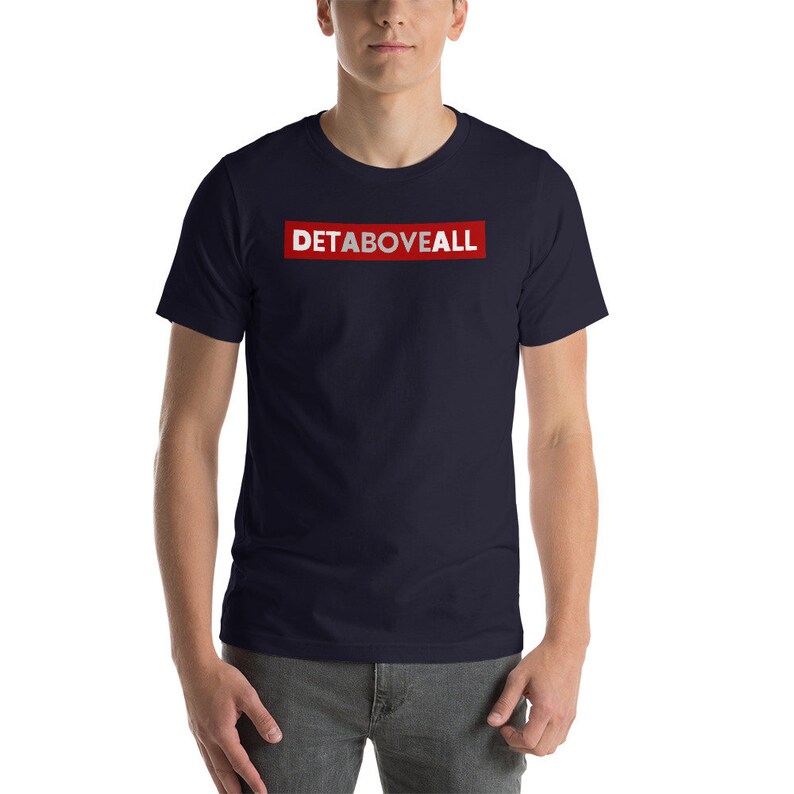 May include: A navy blue t-shirt featuring the text "DETABOVEALL" in white within a red rectangular box. The shirt has a crew neck and short sleeves.
