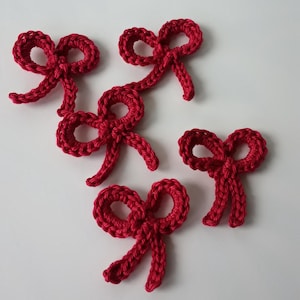 Könnte beinhalten: Fünf handgefertigte, rote Häkel-Schleifen sind auf einer weißen Oberfläche angeordnet. Die Schleifen sind mit sichtbaren Stichen detailliert, und jede hat zwei Schlaufen und zwei fließende Enden. Die Schleifen sind ein festlicher Dekorationsartikel.