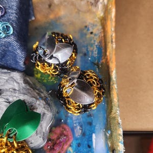 May include: Close-up of two handmade earrings. Each earring features a silver-colored, faceted gem surrounded by a chainmail design in gold and black. The earrings are set against a colorful, textured background.